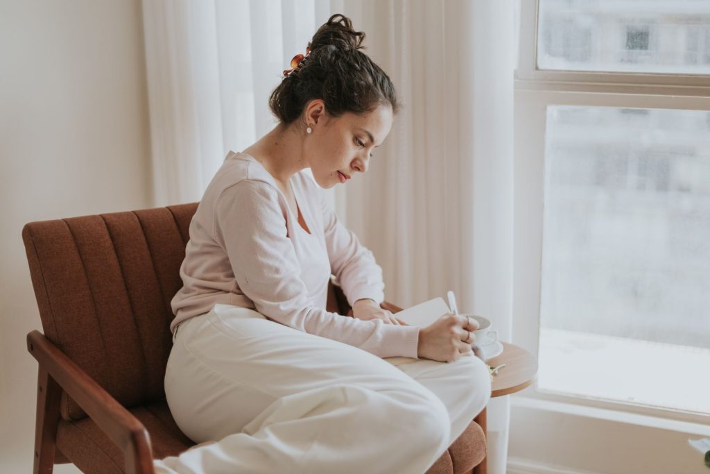 A woman writing in a notebook beside a bright window.
